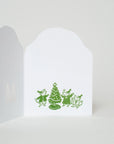 A white greeting card shown open against a light gray background, featuring a green line-art illustration of three children dancing around a Christmas tree.