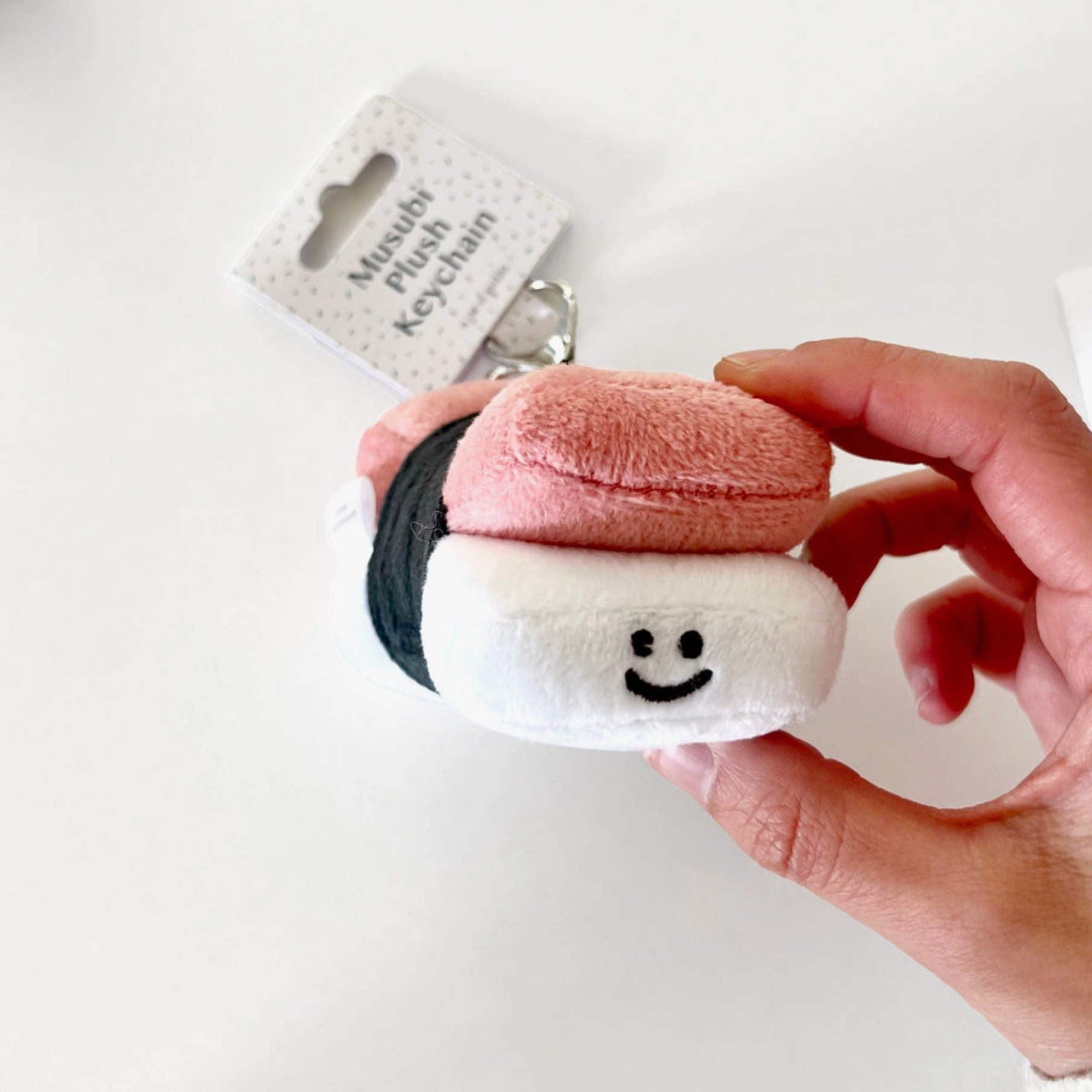 A hand holding a mini plush keychain designed to resemble a spam musubi, with the keychain chain visible and a packaging tag in the background.