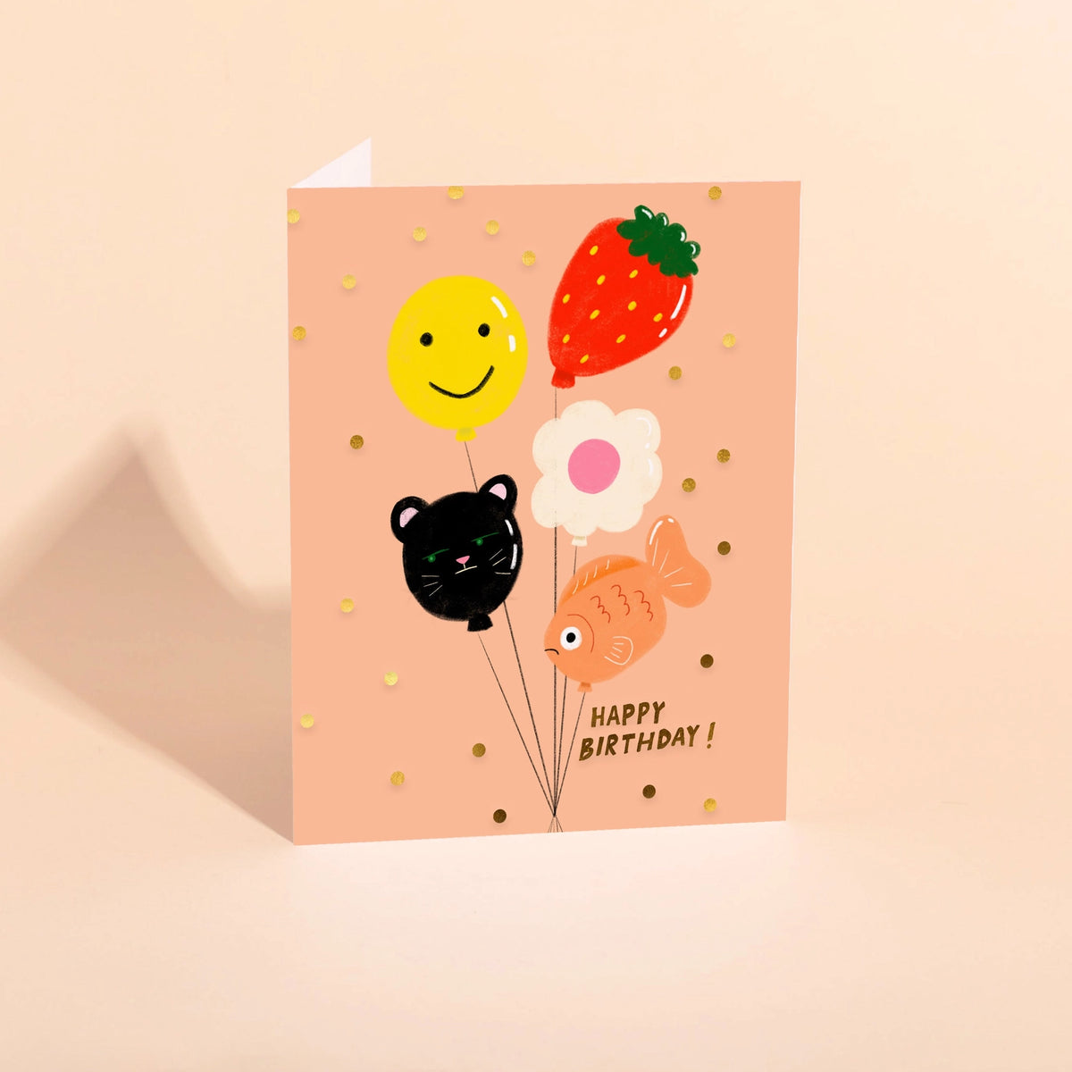 Birthday card with balloons shaped like a smiley face, strawberry, flower, black cat, and goldfish on a pink background.