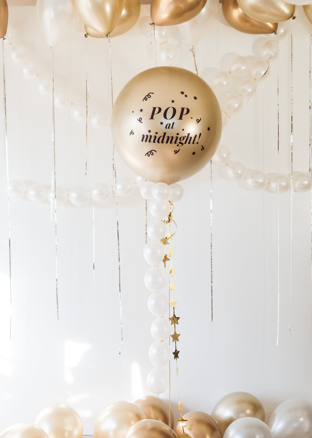 Decorative balloon with 'Pop at midnight!' text hanging among other balloons against a white wooden background.