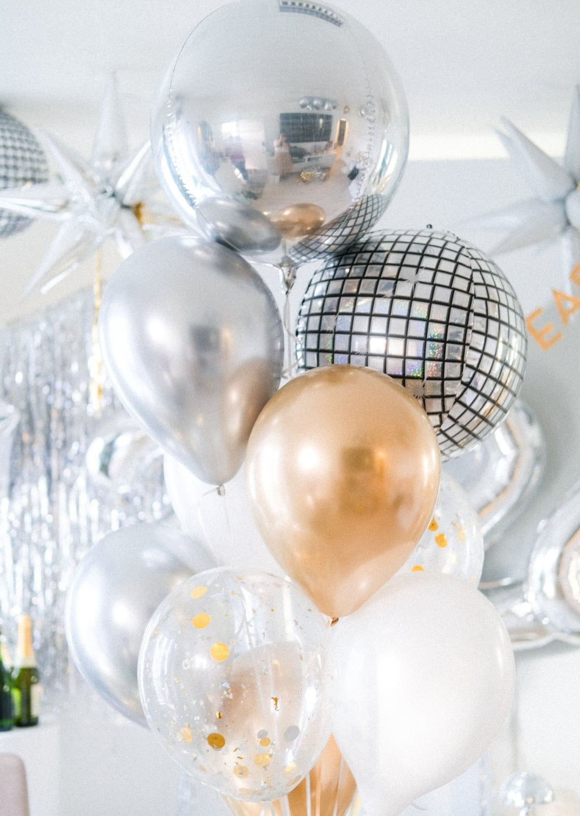 A selection of balloons featuring metallic finishes including a silver mirror orb, with confetti and gold-colored balloons, set against a festive background.