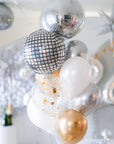A selection of balloons featuring metallic finishes including a silver mirror orb, with confetti and gold-colored balloons, set against a festive background.