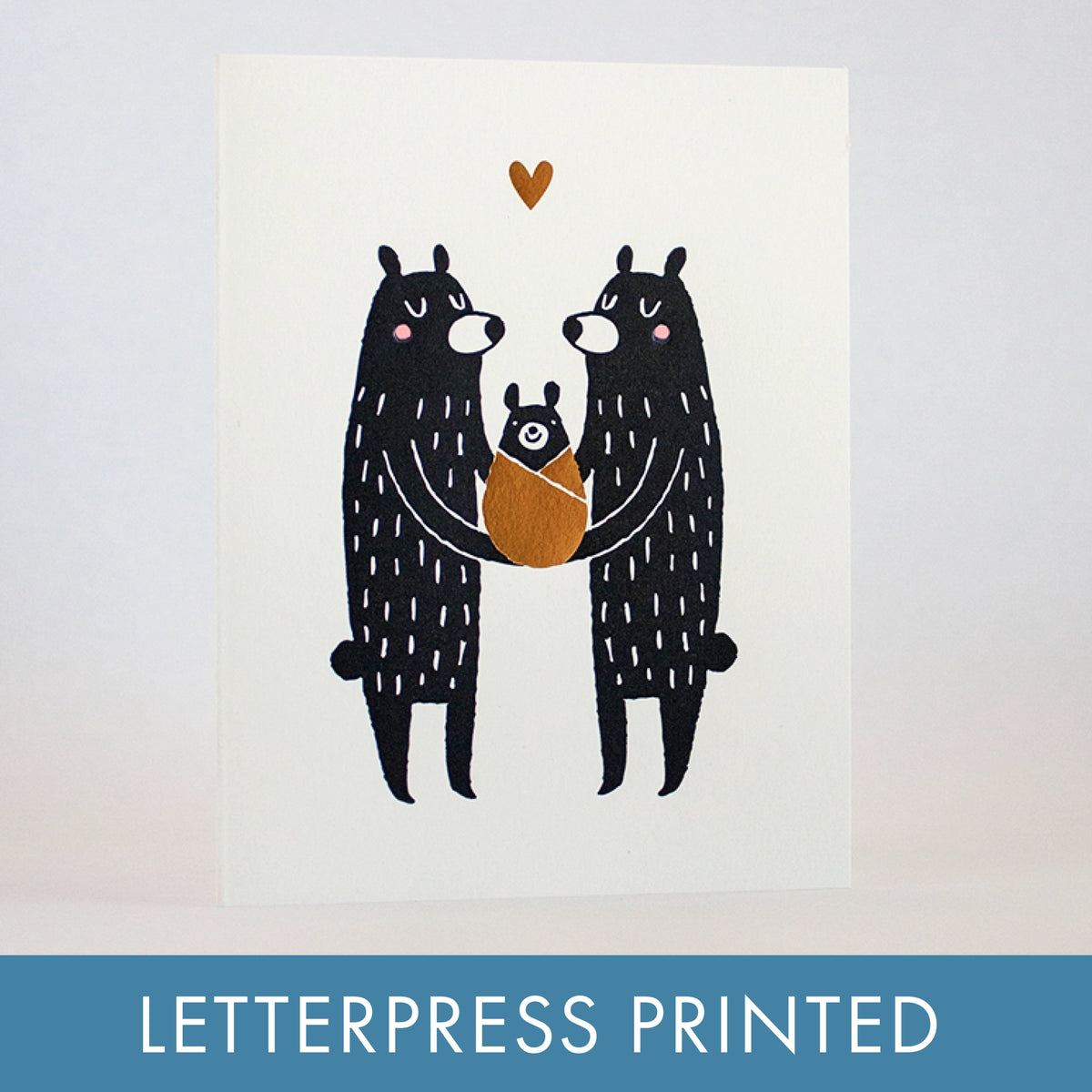 A letterpress printed greeting card featuring a minimalist illustration of two black bears holding a baby bear, with a small gold heart above. The card is displayed against a light gray background with a blue banner at the bottom reading 'LETTERPRESS PRINTED'.