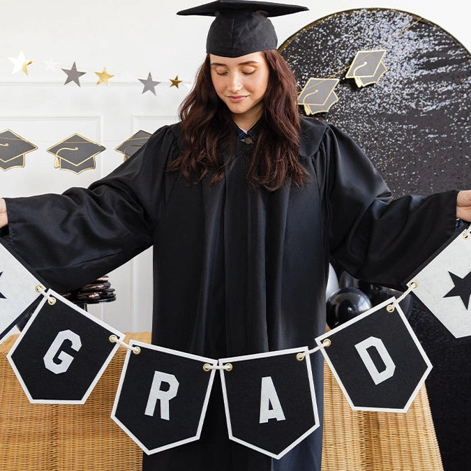 Graduate in cap and gown with 'GRAD' banner against a decorated wall.