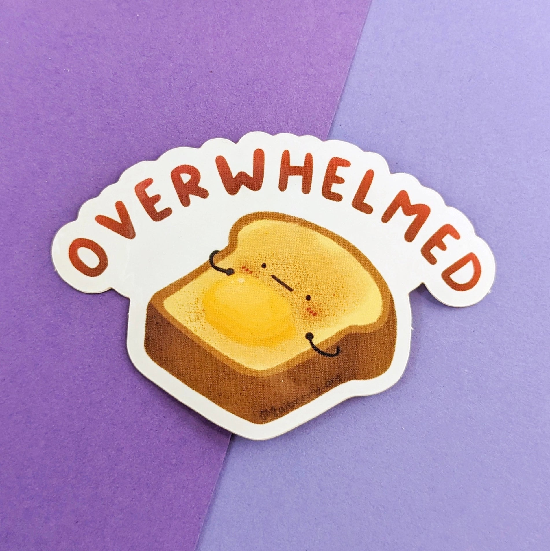 A waterproof glossy sticker depicting a piece of toast with the words 'OVERWHELMED' and a face drawn on it, placed on a two-tone background.