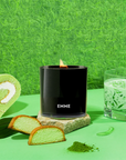 A black candle with a pandan wood wick, placed on a rock, with green background and a glass of milk and a slice of cake in the foreground.