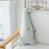 A long-neck plush dinosaur toy in pale mint green with a small party hat and dark embroidered facial features, leaning against a white upholstered chair in a bright indoor setting.