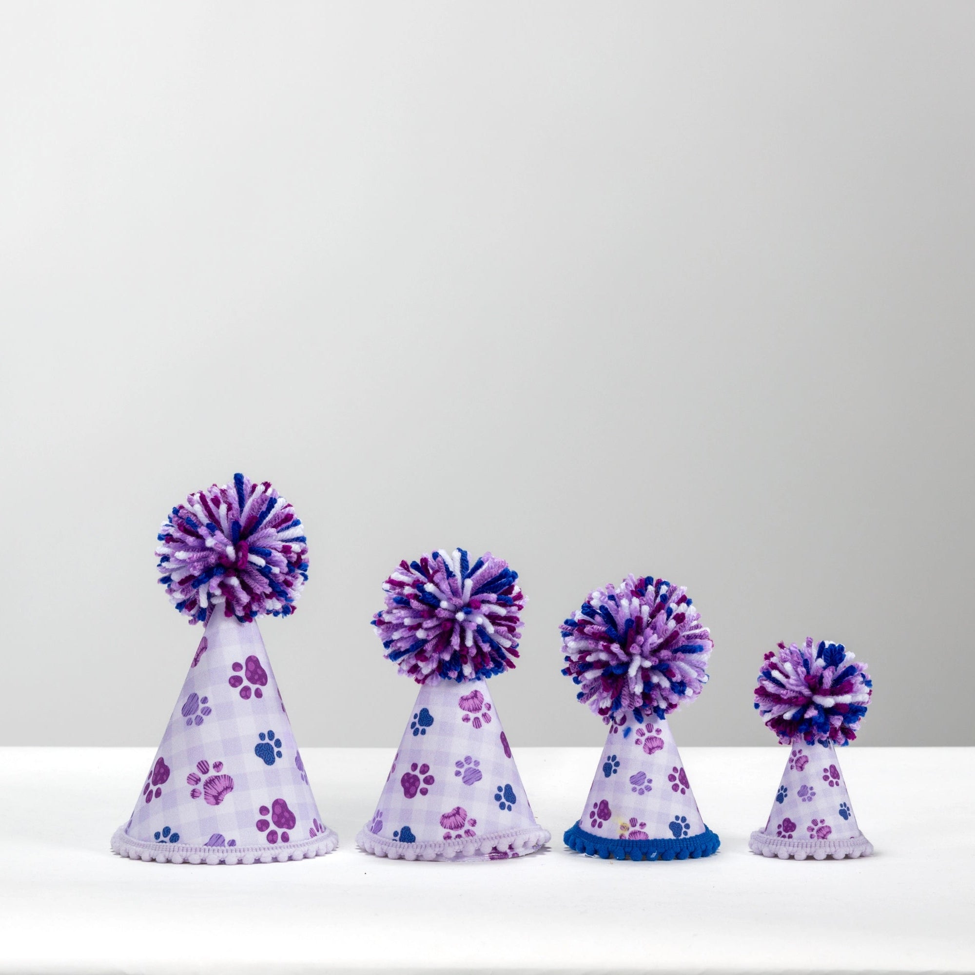 Four small party hats in graduated sizes, decorated with purple paw prints on a white base and topped with purple and blue pom-poms, arranged on a white surface against a light gray background.