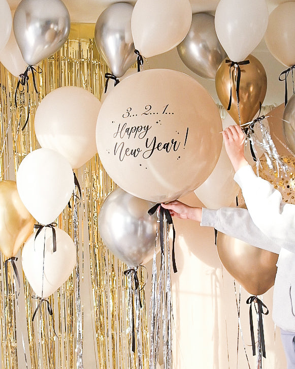 Cream round balloon with black bow and silver metallic tinsel. Text reads "3..2..1.. Happy new Year!" with gold tinsel and cream backdrop. Floating balloon with black ribbon bows surrounding.