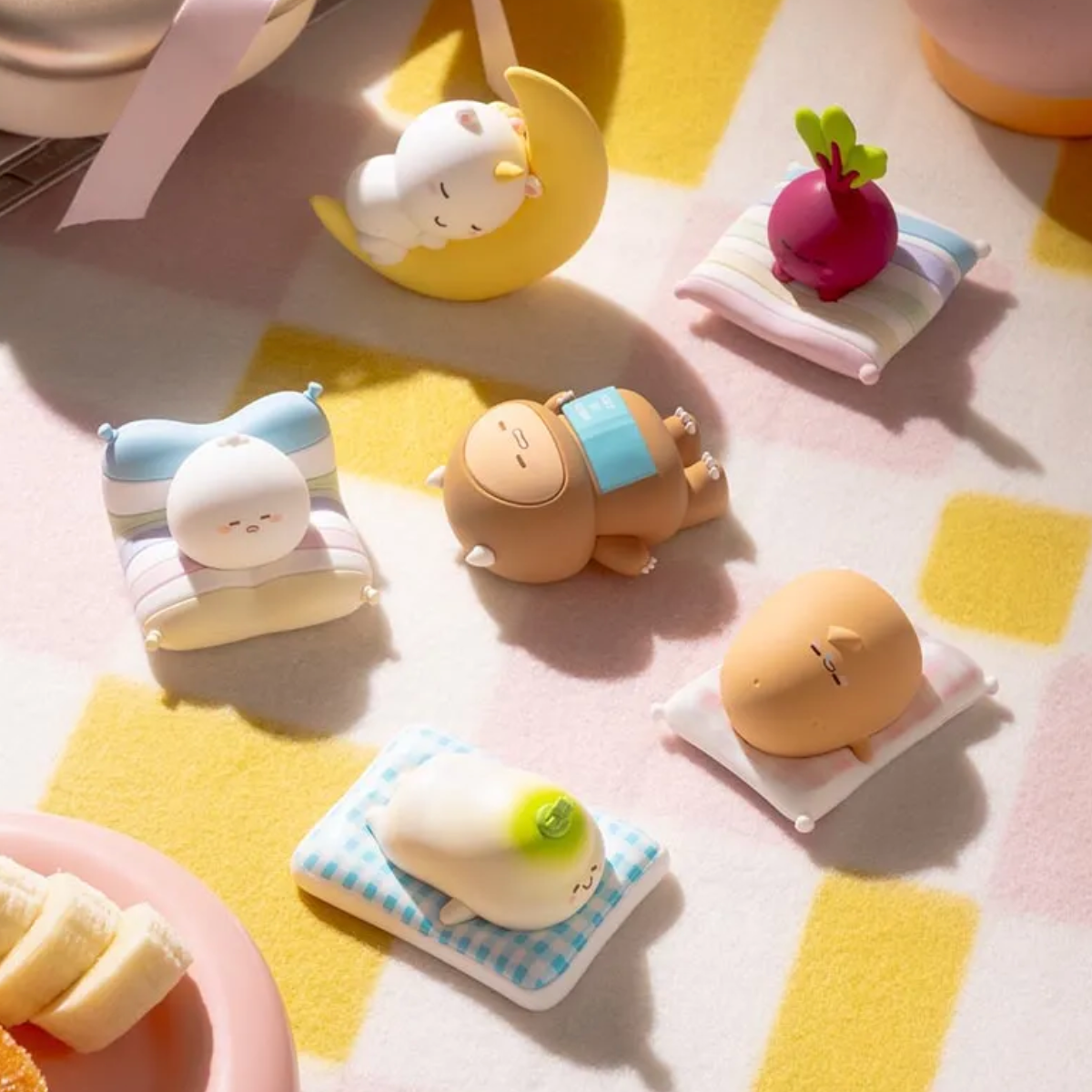 A set of 6 cute, small sleeping character figurines resting on pastel striped pillows on a pink and yellow checkered blanket. 