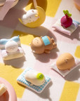 A set of 6 cute, small sleeping character figurines resting on pastel striped pillows on a pink and yellow checkered blanket. 