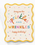 A birthday card with a colorful design, featuring the text 'SPRINKLES AND WRINKLES HAPPY BIRTHDAY!' surrounded by various colorful sprinkles on a light background, also showing a yellow envelope with a square flap.
