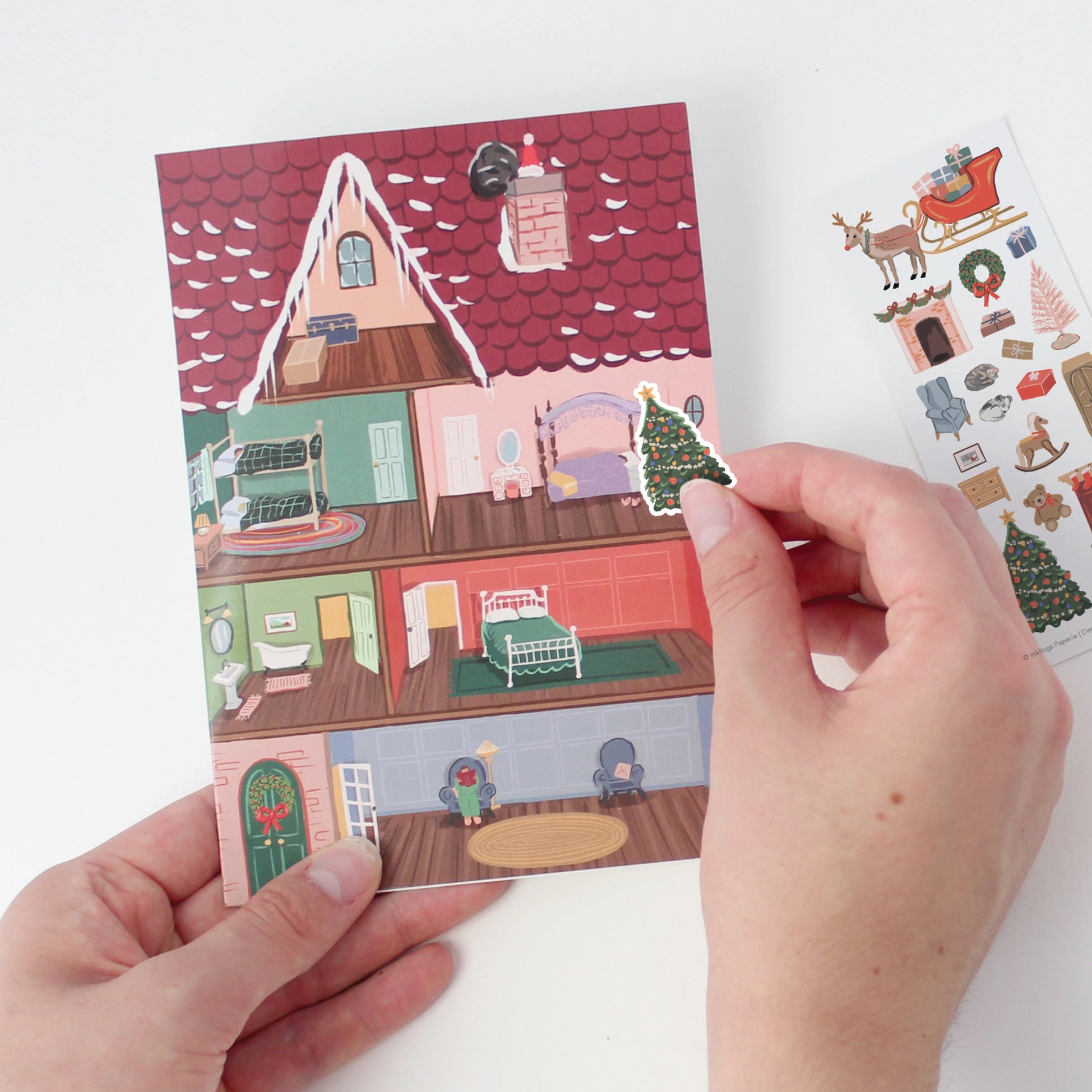 Hand holding a small house-shaped card with a sticker sheet featuring Christmas-themed stickers.