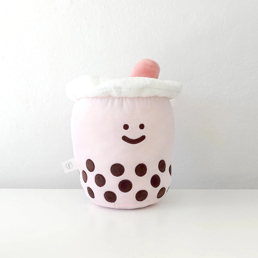 A plush toy shaped like a bubble tea cup, featuring a light pink body with dark brown embroidered polka dots, a white faux-frosted top, and a small pink foam swirl on top, set against a plain white background.