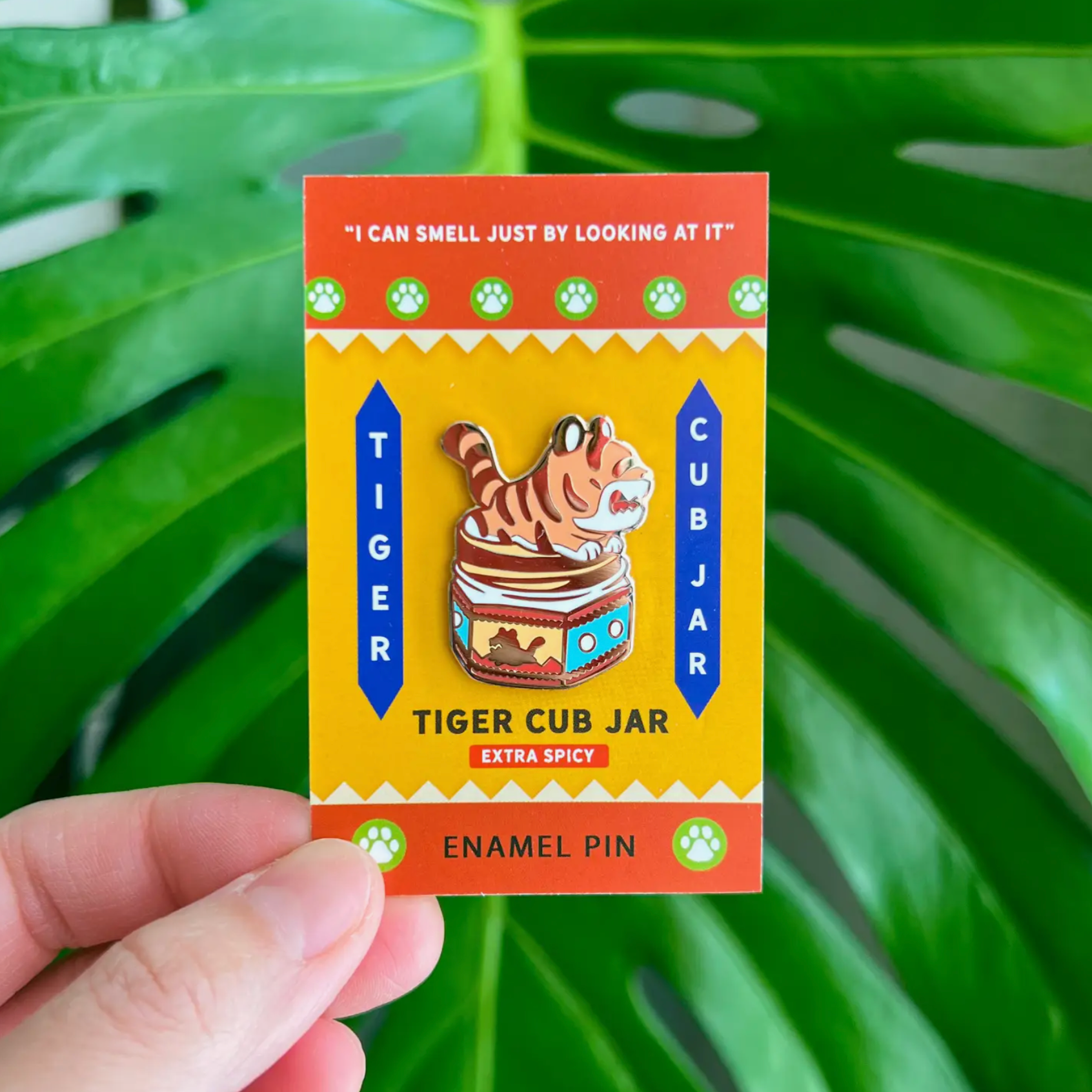 A hand holding a hard enamel pin with a depiction of a tiger inside a jar, alongside a butterfly clutch. The background is green with a leafy pattern.