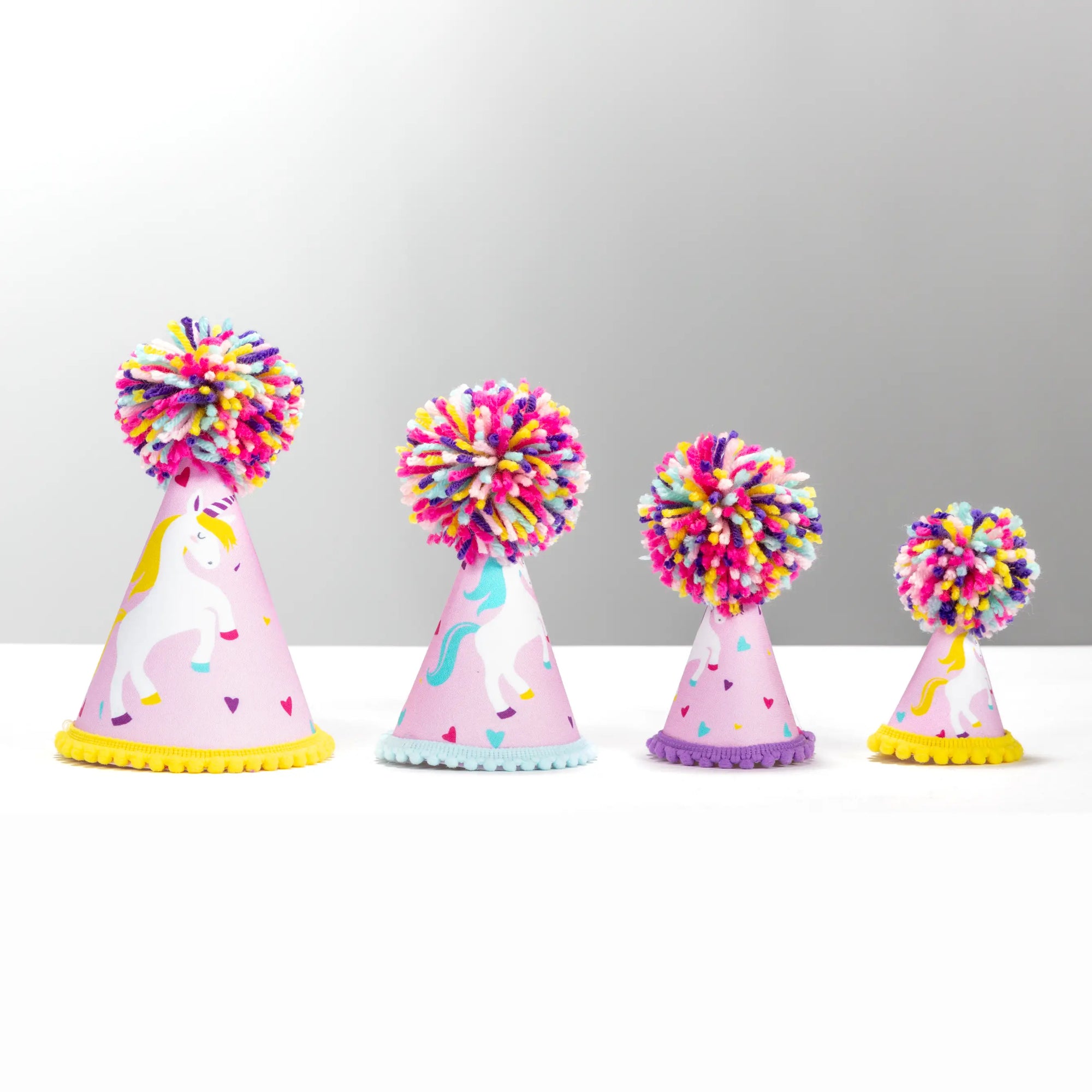 Four unicorn-themed party hats in different sizes lined up on a white surface against a light gray background. Each hat is light pink with a white unicorn illustration, colorful confetti, and a large multicolored pom-pom on top.