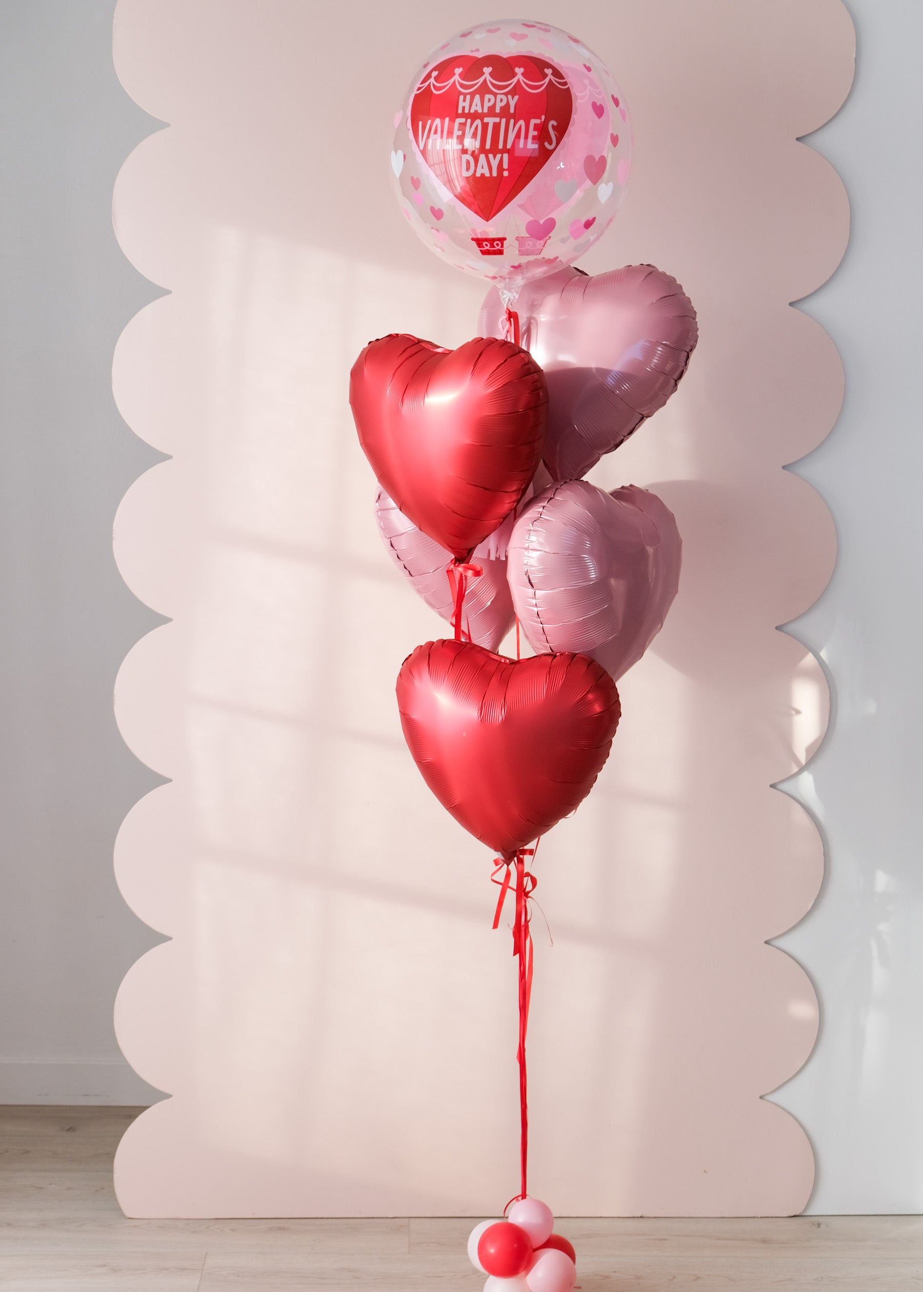 Heart-shaped balloons with a clear 'Happy Valentine's Day' balloon against a decorative pink background.