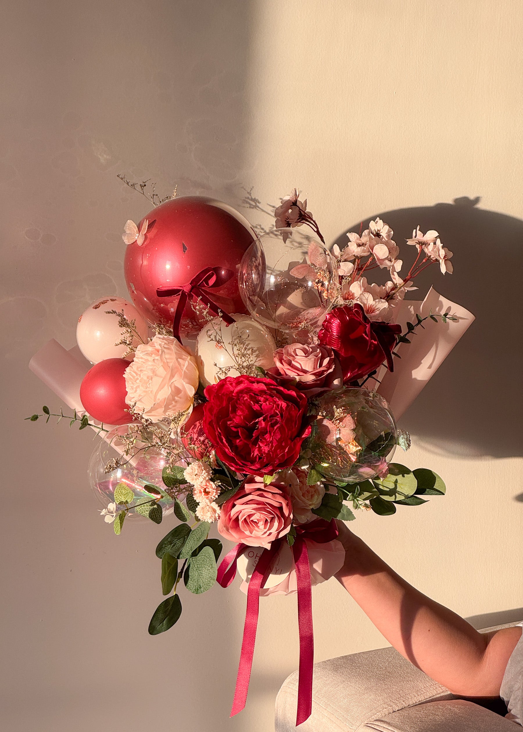 Bouquet of red and pink flowers with balloons held by a person against a neutral background at sunset