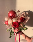 Bouquet of red and pink flowers with balloons held by a person against a neutral background at sunset