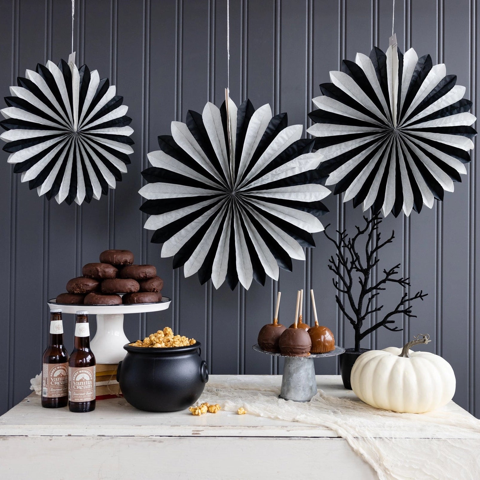 Black and white striped paper fan decorations hanging on a dark gray paneled wall above a styled Halloween dessert table with cookies, candy apples, a cauldron, and a white pumpkin.