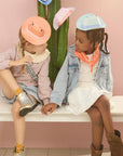 Two children sitting on a bench with colorful hats and scarves, against a pink wall.