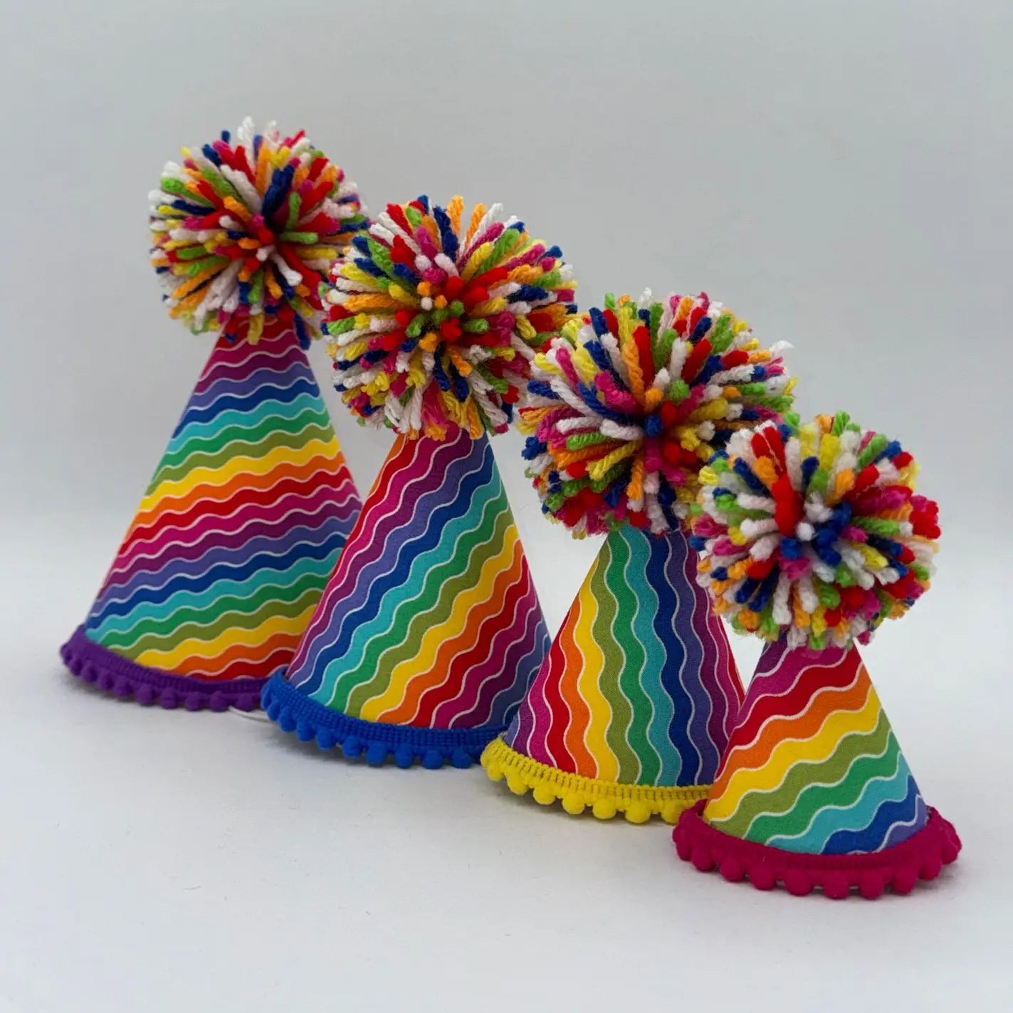 Four colorful party hats with rainbow wavy stripes and fluffy multicolor pom-poms, arranged on a light gray background.