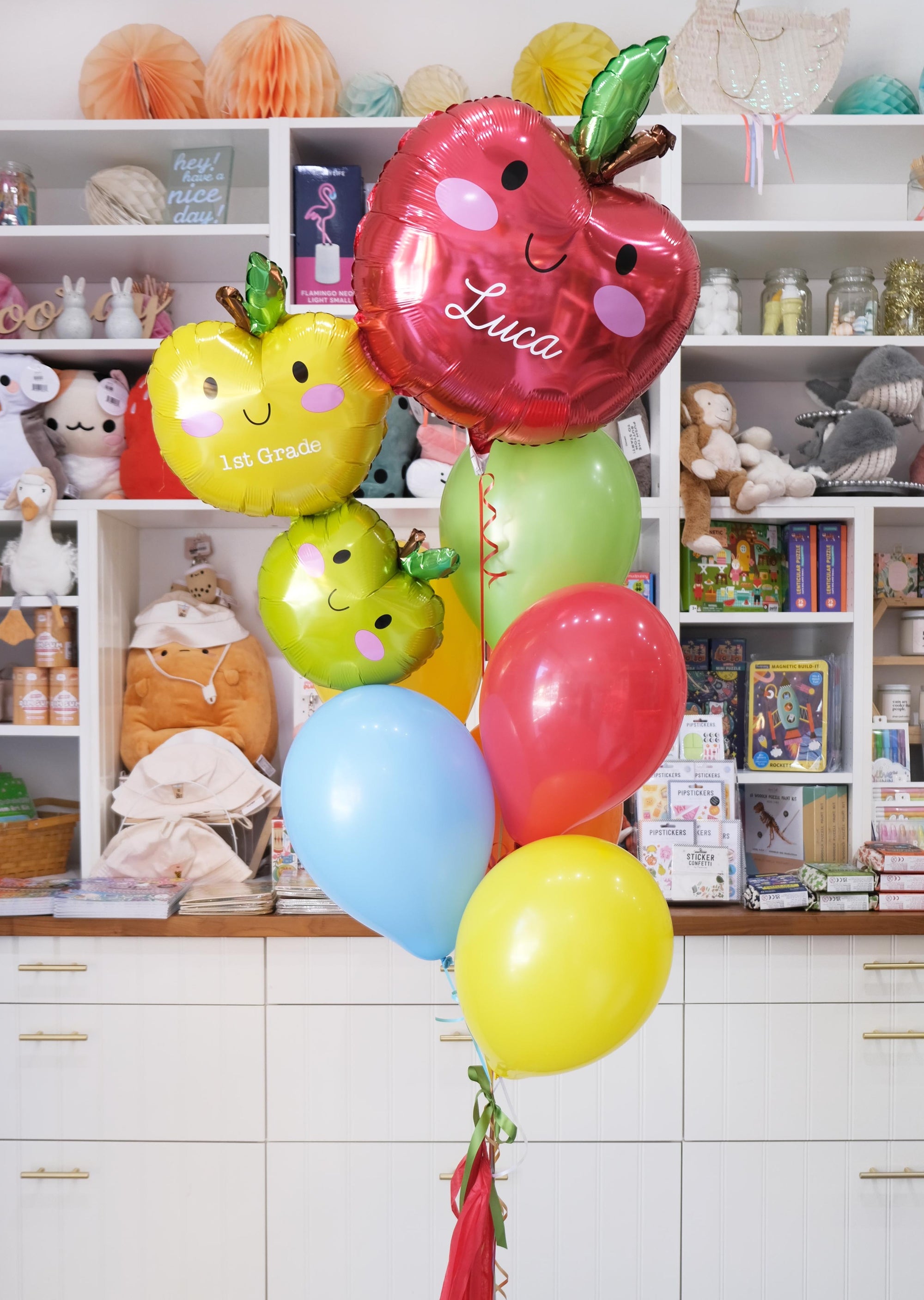 A set of colorful balloons designed to look like apples with the name 'Luca' and the grade '1st Grade' printed on them, along with a handmade tassel.