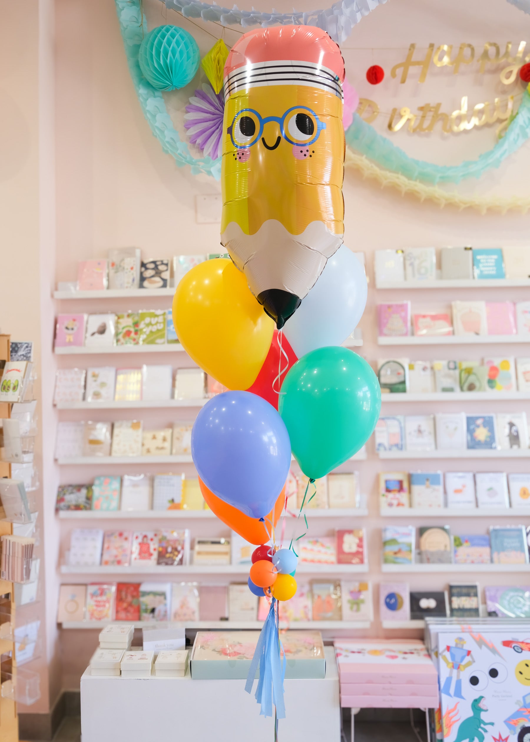 a balloon bouquet featuring a pencil balloon in front of a wall of greeting cards