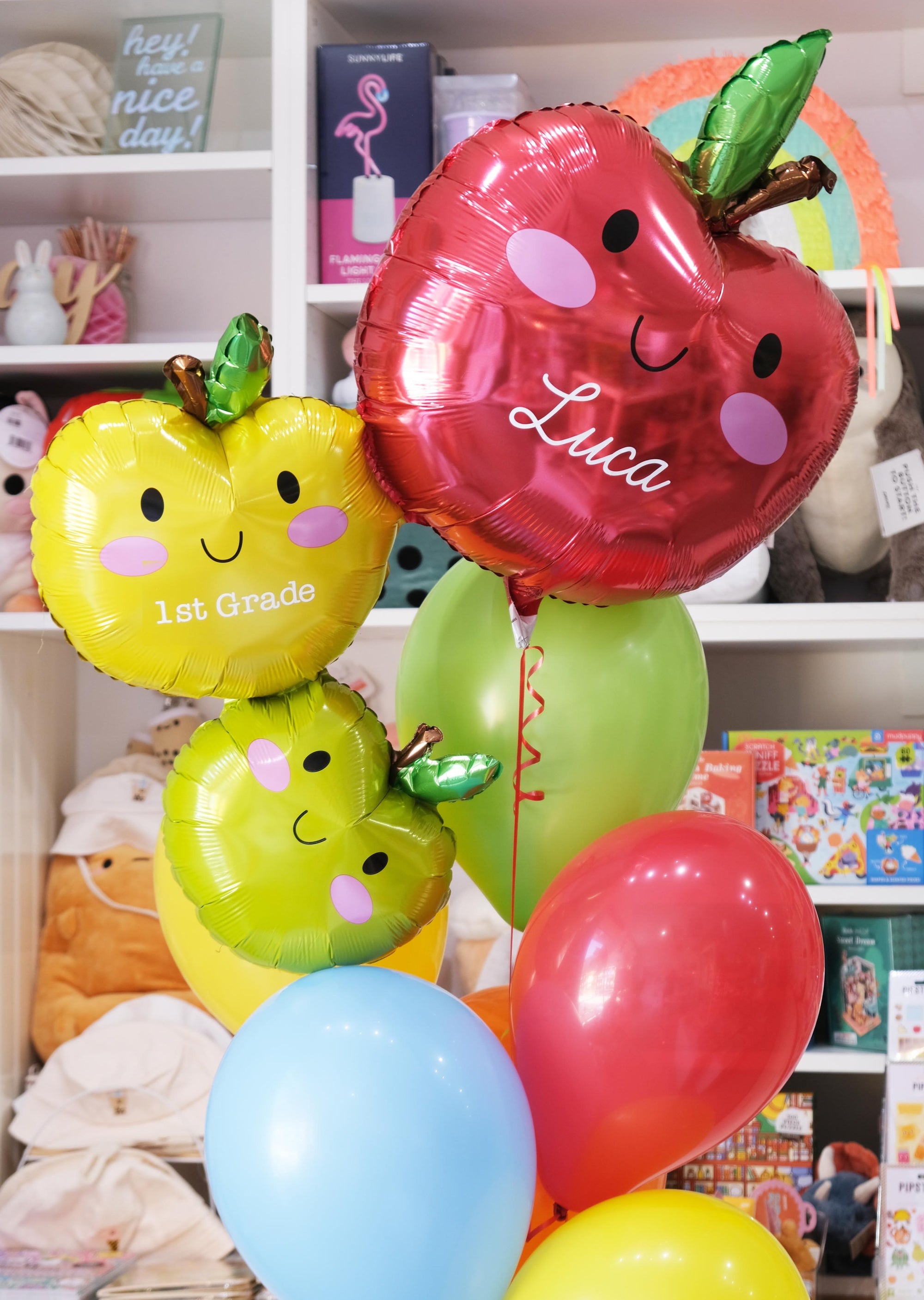 A set of colorful balloons designed to look like apples with the name 'Luca' and the grade '1st Grade' printed on them, along with a handmade tassel.