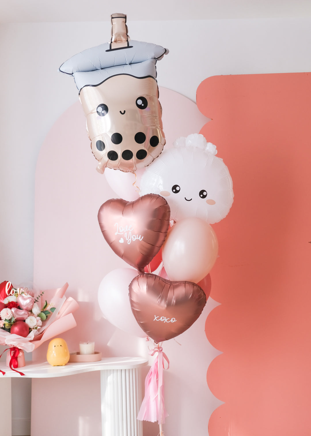 A balloon bouquet featuring a boba bubble tea character balloon, a dumpling with a heart face balloon, and a variety of other balloons in pink, red, and white colors with 'love you' and 'xoxo' decals.