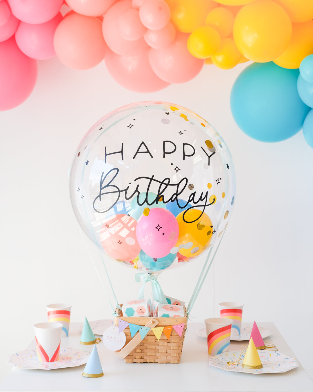 A decorative hot air balloon basket with 'Happy Birthday' text, containing cupcakes and adorned with colorful balloons and confetti inside. A cluster of balloons in rainbow colors decoarte above the table.