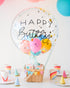 A decorative hot air balloon basket with 'Happy Birthday' text, containing cupcakes and adorned with colorful balloons and confetti inside.