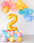An indoor balloon garland featuring a large gold number 2 amidst a mix of colorful balloons in shades of pink, yellow, blue, and teal, with a 'Happy Birthday' message on the top large balloon.