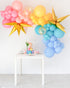 A balloon garland featuring a variety of colored balloons including pink, yellow, blue, and multicolored starburst and confetti balloons, displayed indoors next to a table with a decorative item.