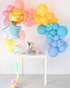 An assortment of colorful balloons in pink, yellow, blue, and gold, arranged in a garland style with a table and chairs in the foreground.
