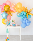 An assortment of colorful balloons in pink, yellow, blue, and gold, arranged in a garland style with a table and tableware in the foreground.
