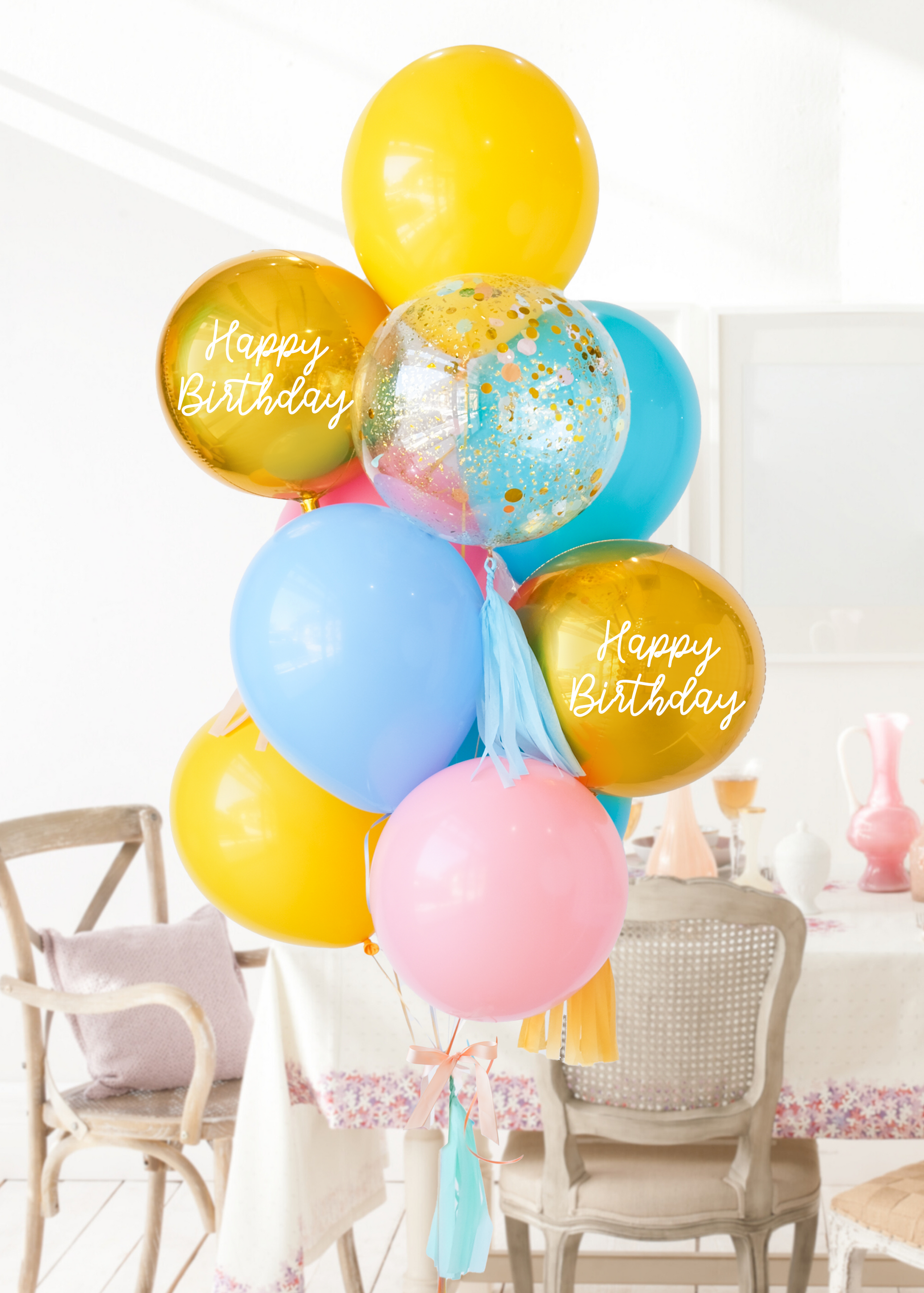 A balloon bouquet with a mix of solid colored balloons in blue, yellow, pink, and green, along with two metallic orb balloons, one with 'Happy Birthday' text, and a clear confetti balloon. There is a tassel and a ribbon bow on the arrangement. Bouquet is in front of a dining table and chairs