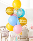 A balloon bouquet with a mix of solid colored balloons in blue, yellow, pink, and green, along with two metallic orb balloons, one with 'Happy Birthday' text, and a clear confetti balloon. There is a tassel and a ribbon bow on the arrangement. Bouquet is in front of a dining table and chairs