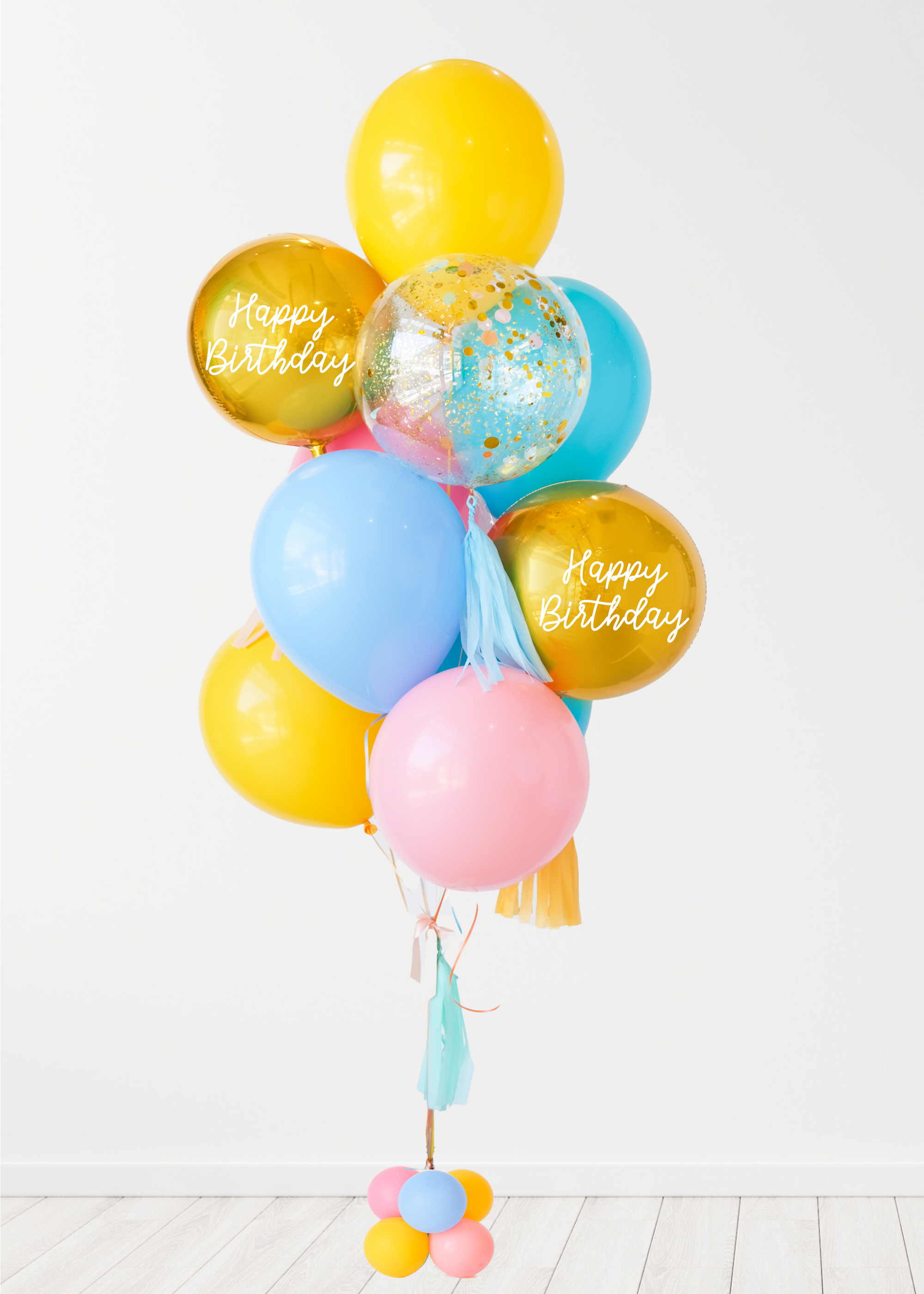 A balloon bouquet with a mix of solid colored balloons in blue, yellow, pink, and green, along with two metallic orb balloons, one with 'Happy Birthday' text, and a clear confetti balloon. There is a tassel and a ribbon bow on the arrangement. White wall background.
