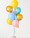 A balloon bouquet with a mix of solid colored balloons in blue, yellow, pink, and green, along with two metallic orb balloons, one with 'Happy Birthday' text, and a clear confetti balloon. There is a tassel and a ribbon bow on the arrangement. White wall background.