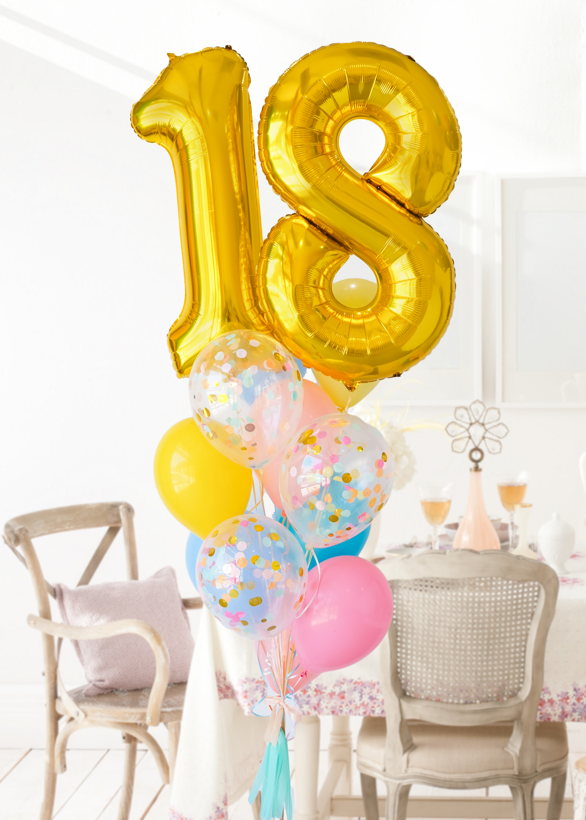 A balloon bouquet featuring a large gold number 18 balloon, with smaller multicolored balloons including pink, blue, and yellow, all adorned with confetti patterns. The balloons are attached to a ribbon bow and tassel. Bouqet is in a dining room setting.