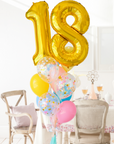 A balloon bouquet featuring a large gold number 18 balloon, with smaller multicolored balloons including pink, blue, and yellow, all adorned with confetti patterns. The balloons are attached to a ribbon bow and tassel. Bouqet is in a dining room setting.