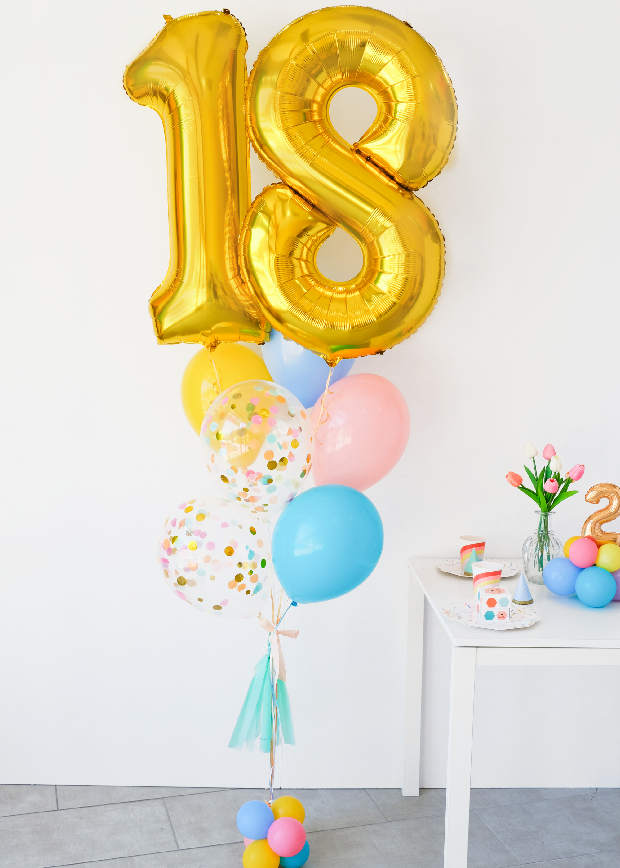 A balloon bouquet featuring a large gold number 18 balloon, with smaller multicolored balloons including pink, blue, and yellow, all adorned with confetti patterns. The balloons are attached to a ribbon bow and tassel. A snippet of a tabel with tulips and small balloon centerpiece to the right.