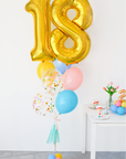 A balloon bouquet featuring a large gold number 18 balloon, with smaller multicolored balloons including pink, blue, and yellow, all adorned with confetti patterns. The balloons are attached to a ribbon bow and tassel. A snippet of a tabel with tulips and small balloon centerpiece to the right.