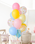 A bouquet of balloons in shades of blue, pink, yellow, and multicolor with a confetti pattern. Bouquet is in a dining room setting