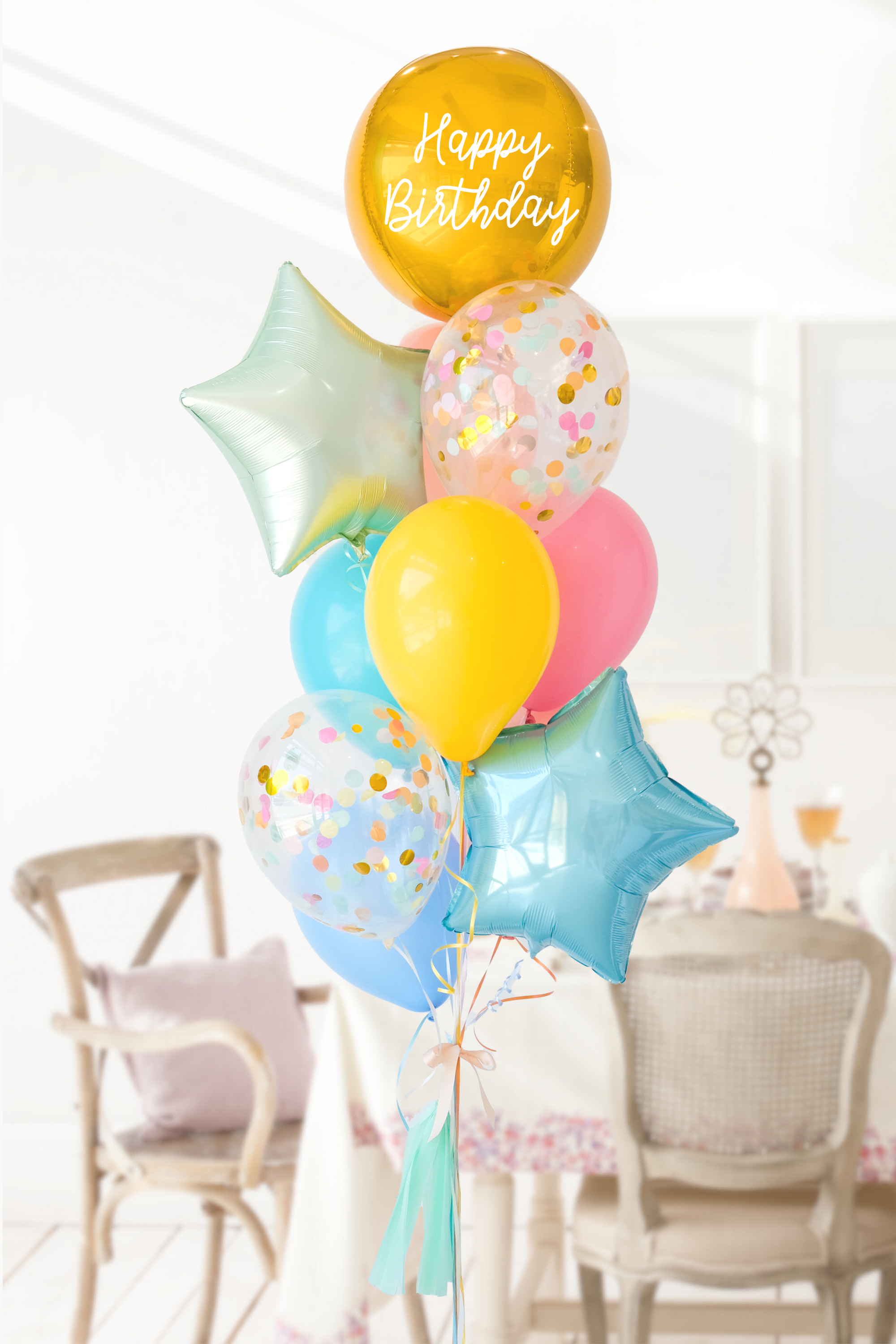 A balloon bouquet featuring a round gold orb balloon, with smaller multicolored balloons including pink, blue, and yellow, all adorned with confetti clear balloons and stars. The balloons are attached to a ribbon bow and tassel. Bouquet is in a dining room setting.