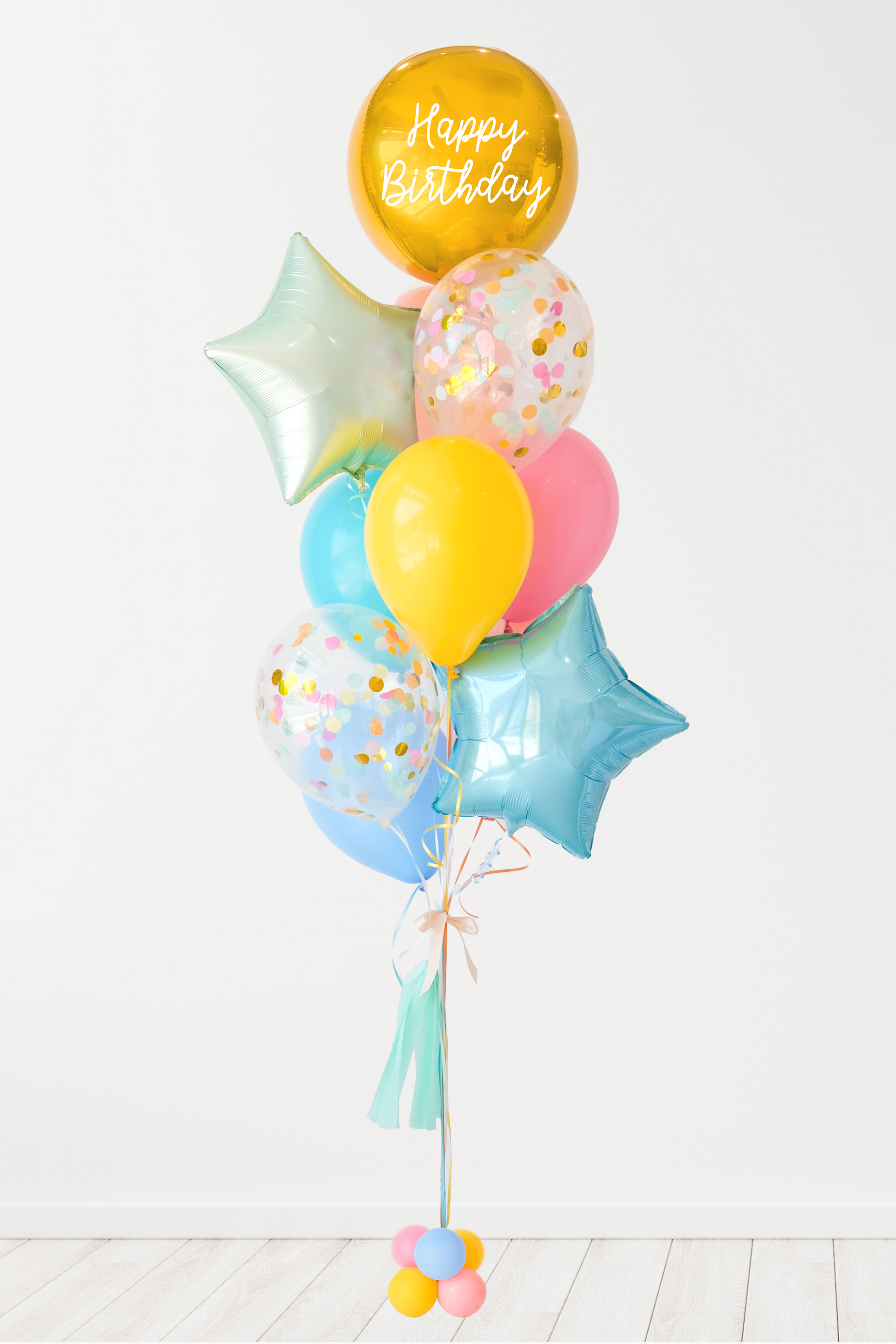 A balloon bouquet featuring a round gold orb balloon, with smaller multicolored balloons including pink, blue, and yellow, all adorned with confetti clear balloons and stars. The balloons are attached to a ribbon bow and tassel. White background.