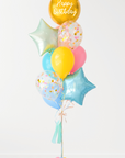 A balloon bouquet featuring a round gold orb balloon, with smaller multicolored balloons including pink, blue, and yellow, all adorned with confetti clear balloons and stars. The balloons are attached to a ribbon bow and tassel. White background.