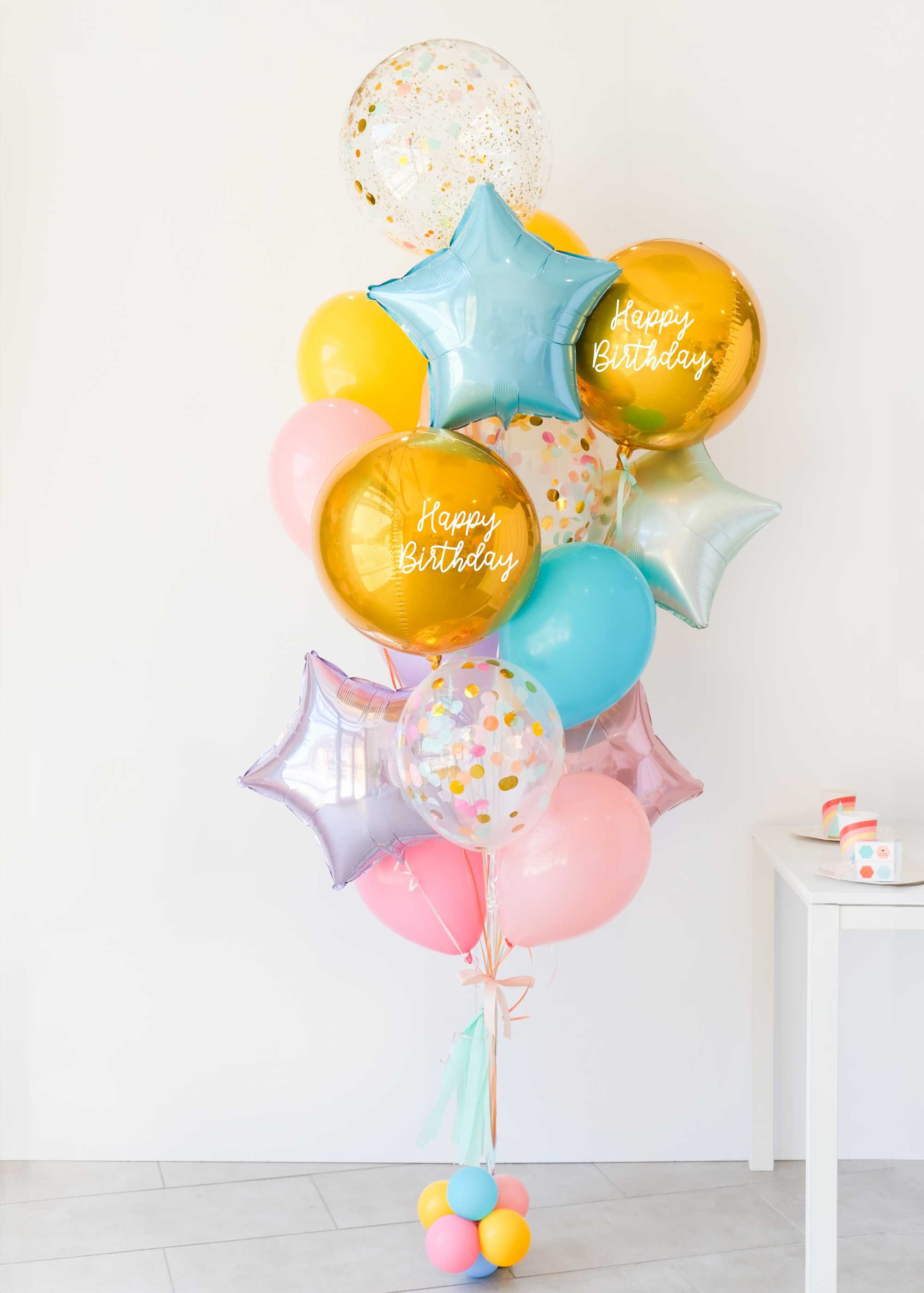 A balloon bouquet featuring two round gold orb balloon, with smaller multicolored balloons including pink, blue, and yellow, all adorned with confetti clear balloons and stars. The balloons are attached to a ribbon bow and tassel.