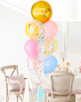 A balloon bouquet with a mix of solid colored balloons in blue, yellow, pink, and green, along with one metallic orb balloon with 'Happy Birthday' text, and a clear confetti balloon. There is a tassel and a ribbon bow on the arrangement. Bouquet is in a dining room setting.